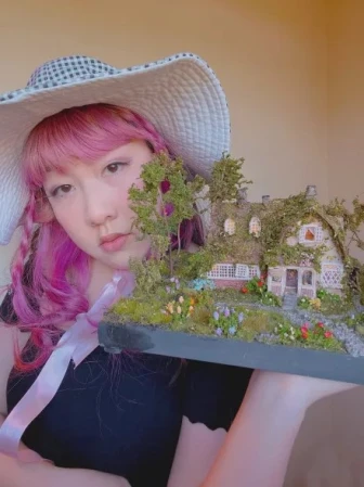 Gloria holding up a miniature house she made. It's awesome; highy detailed. It has grass and flowers and trees in the front yard and there's moss and branches growing over the roof of the cottage-like house. It has a cute porch.