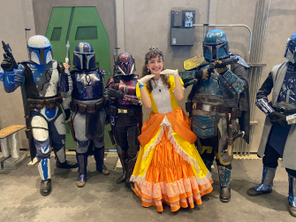 Annie Wiebe, dressed as Daisy -- with tiny crown, white gloves, and a yellow and orange dress -- smiling -- surrounded by people in Star Wars Mandolorian gear.