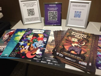 Various Winnipeg Game Jam posters laid out on a table. This year's poster has an adorable white cat in a circus ringmaster shirt swinging on a cord connected to a gaming controller. Behind the posters are QR codes for New Media Manitoba and IDM.