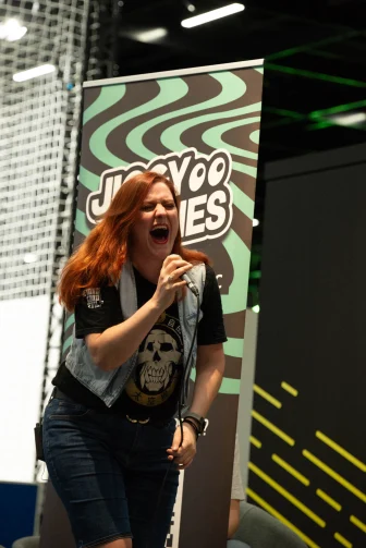 PHOTO CREDIT: kingsmanfotos. Photo is Jayhan screaming into a microphone at Gamescom 2023. She's got a punk look: denim sleeveless jacket and a black shirt with a skull on it and jean shorts. A poster for - I think - Jiggy Tunes is in the background