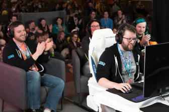 Supplied photo of Amyrlinn at AGDQ. He's in a white gaming chair, hand on the mouse, looking at a computer monitor. Behind him are his commentators and adoring fans. (photo from GDQ)